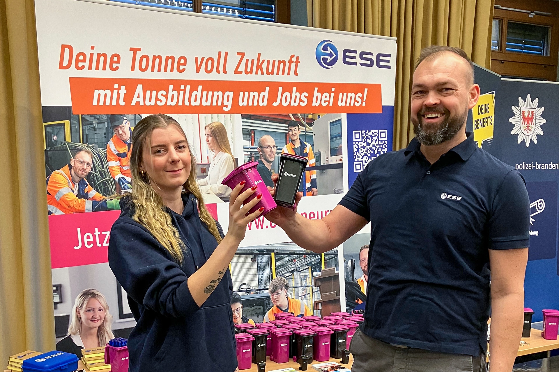 Stephan Schillkowski von ESE (rechts) steht mit Annabelle (links), ESE-Auszubildende im Büromanagement, am ESE Stand auf der Ausbildungsmesse 2025 im OSZ Neuruppin. Beide halten die Giveaways in Form kleiner Mülltonnen in pink und schwarz in die Kamera und Lächeln. Hinter Ihnen sieht amn den Infostand mit weiteren Giveaways und Infomaterialien zu den verschiedenen Ausbildungsberufen bei ESE.