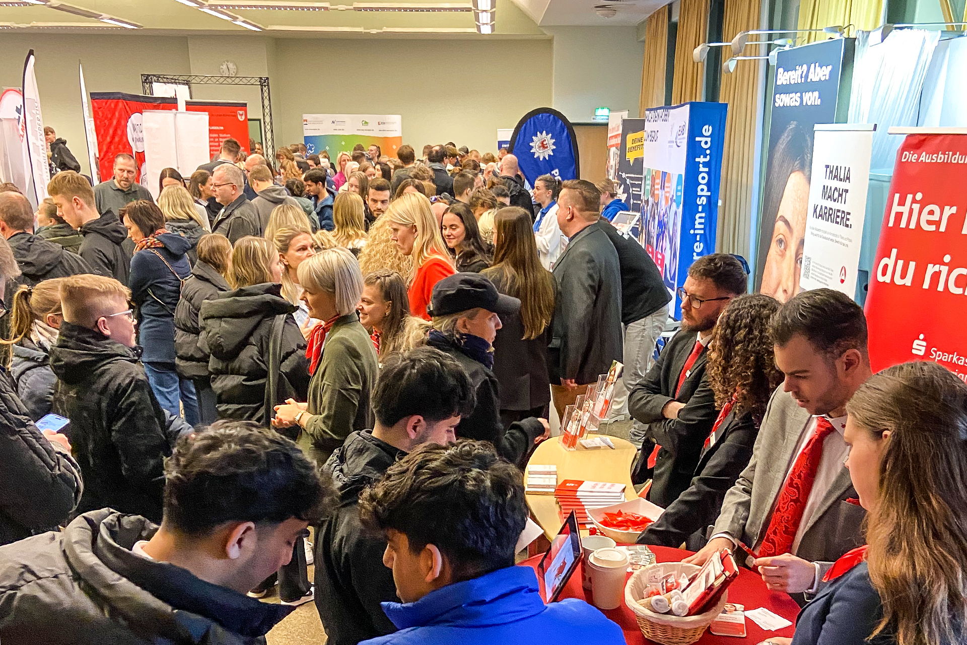Raumfoto mit großem Besucherandrang auf der Ausbildungsmesse 2025 im OSZ Neuruppin. An verschiedenen Ständen stehen Jugendliche udn Familien und sprechen mit den jeweiligen Firmenvertretern.